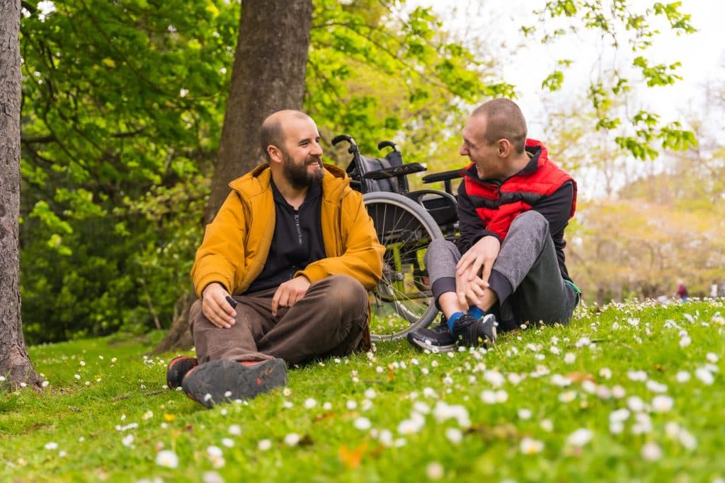 Two people laughing together outdoors in a park, with a wheelchair nearby — inclusive community support